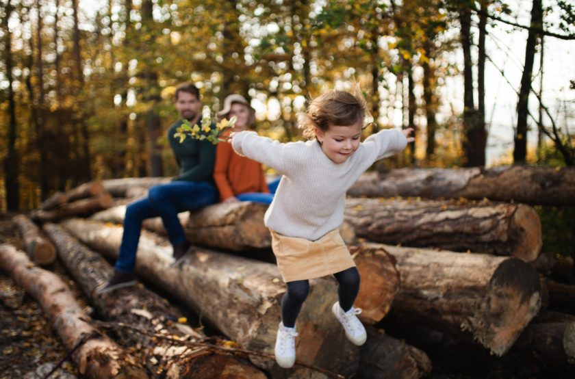 little girl jumping off big logs in forest while respectful parents watch