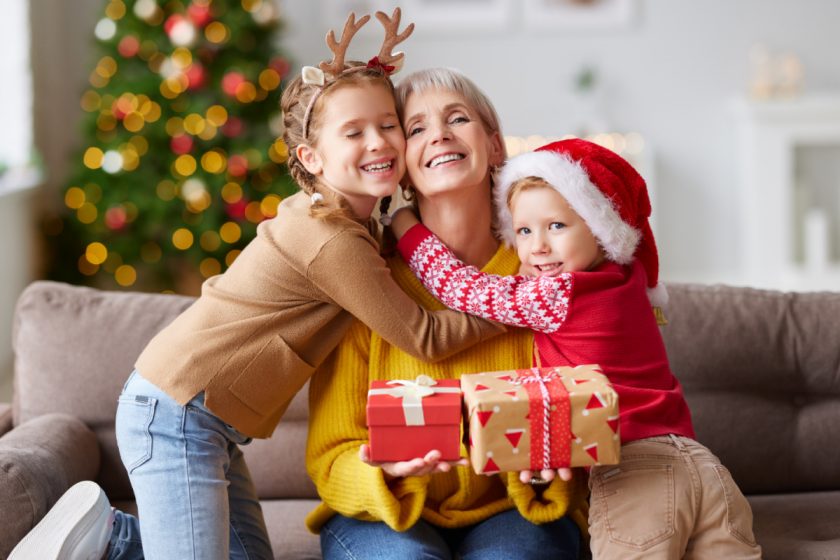 Surprised elderly grandmother with Christmas gift box and looking excited while hugging with grandchildren at home