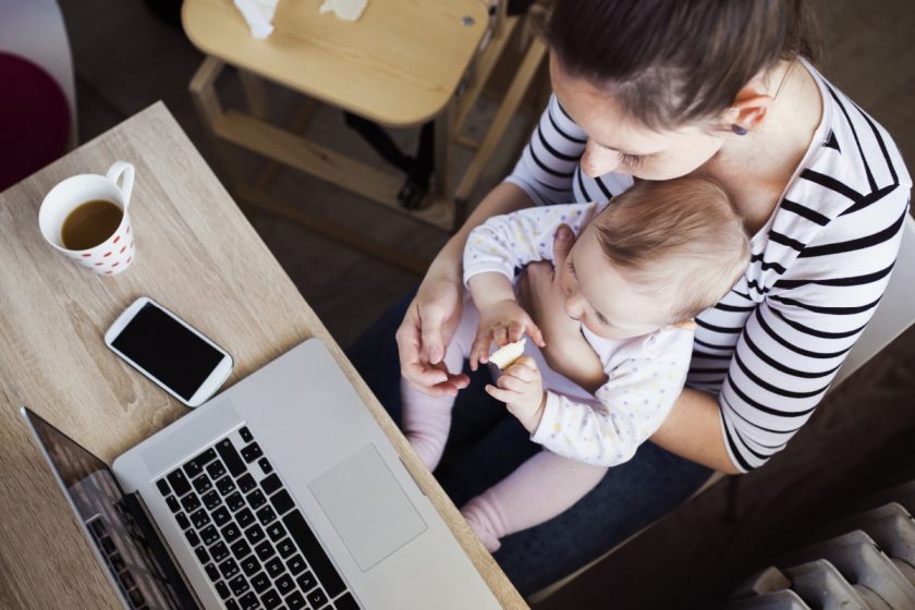 work from home mom holding baby on her lap while trying to get work done, laptop, phone and coffee on desk
