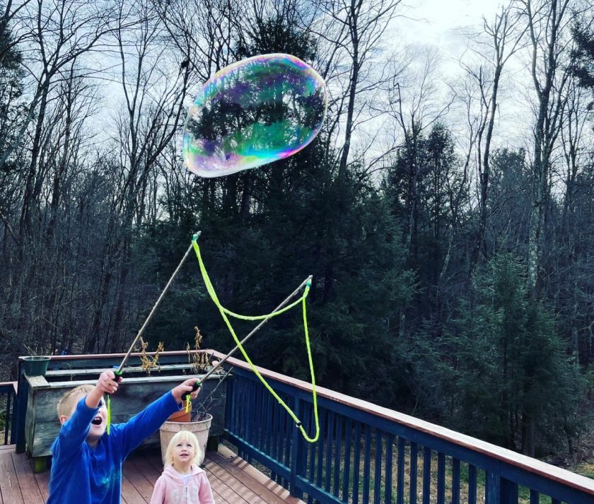two unschooled children making gigantic bubbles on deck with trees in background