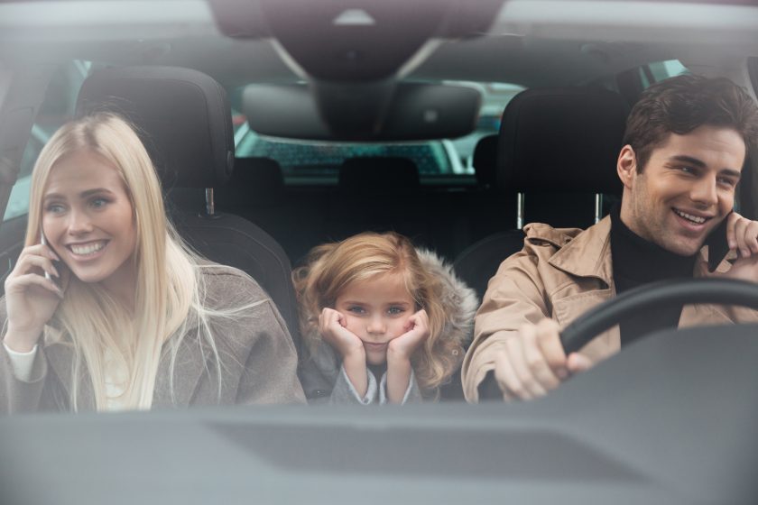 Sad little girl sitting in car while her mother and father talking by mobile phones.