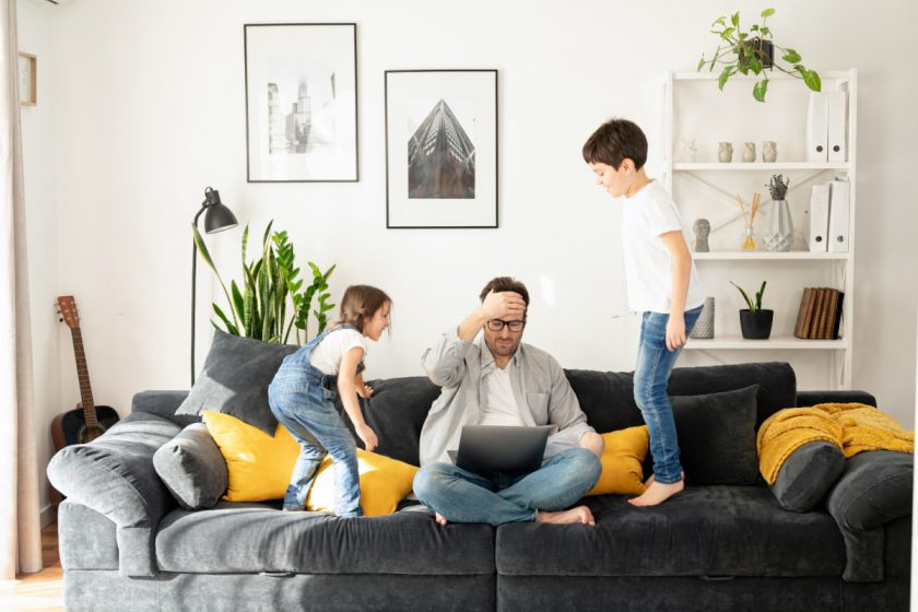 tired dad trying to work with laptop on couch, surrounded by two jumping children