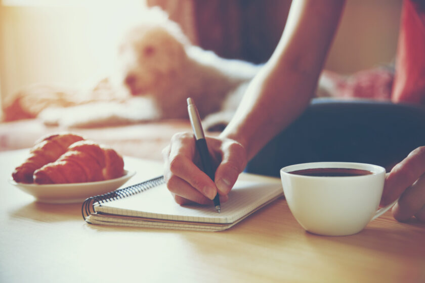 woman's hand with pen writing on notebook during her minimalist morning routine with coffee and croissants