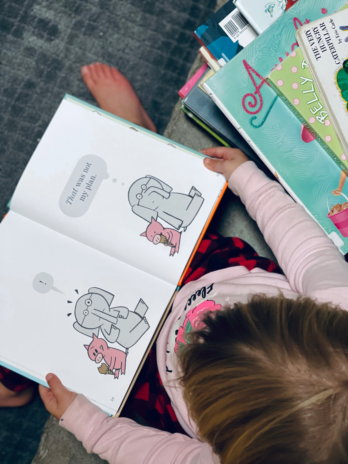 3-year-old sitting on mantle with Elephant and Piggie book on her lap and stack of books beside her