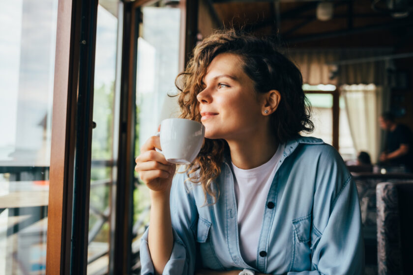 Smiling calm young woman drinking coffee and dreamily looking out the window sitting at table in restaurant.