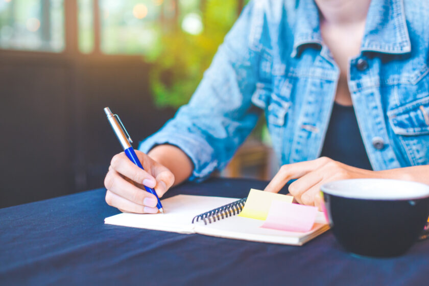 woman writing down her morning routine in a notebook with post-its, while drinking morning coffee