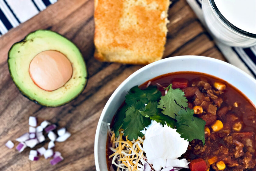 sweet and healthy turkey chili in white bowl on cutting board with avocado, cornbread and chopped red onion