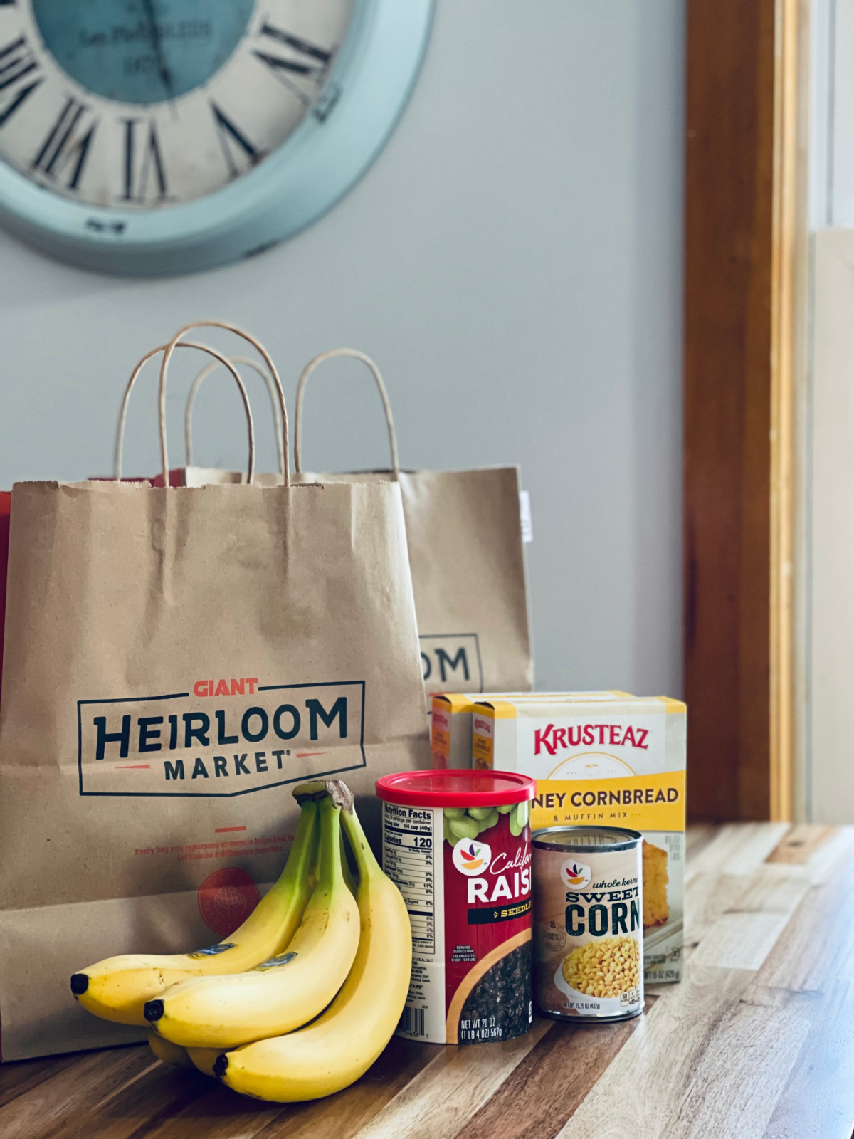 Giant grocery bags and grocery items sitting on kitchen table with clock in background
