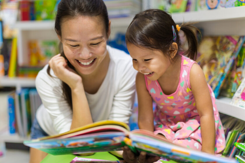 young Asian mom smiling, enjoying reading to her young daughter.