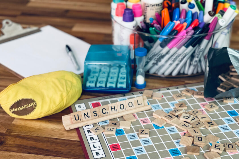 letter tiles spelling "unschool" on top of Scrabble board, with Boggle, bananagrams, clipboard and markers in background