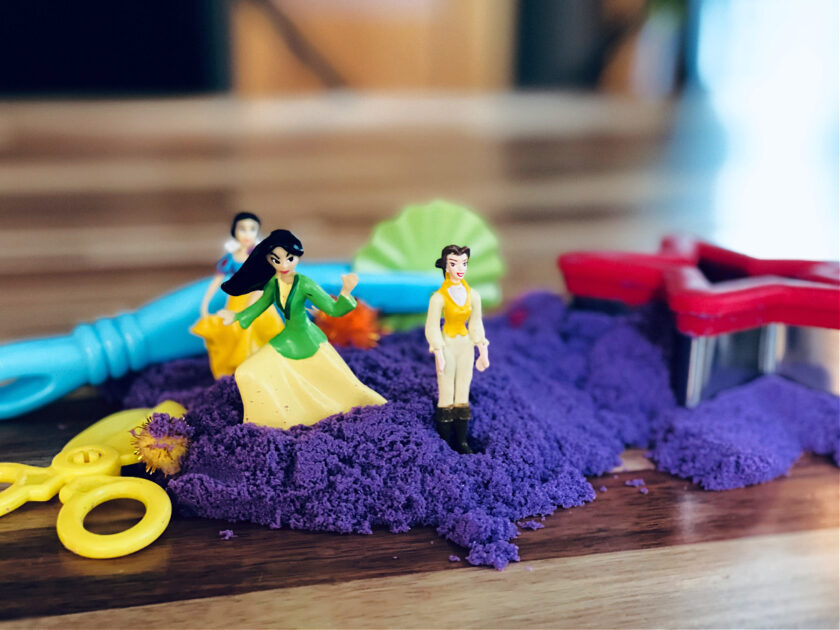 Disney princess figurines and playdough tools in purple kinetic sand on kitchen table.