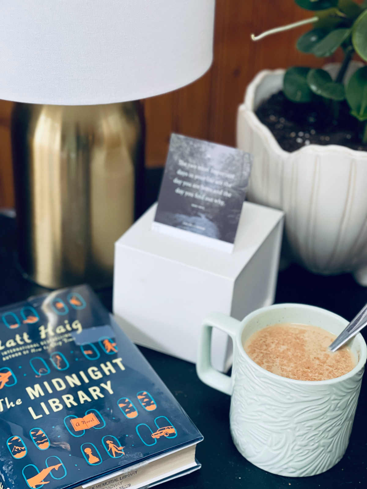 coffee and book The Midnight Library sitting on table near lamp, plant and calendar quote cards.