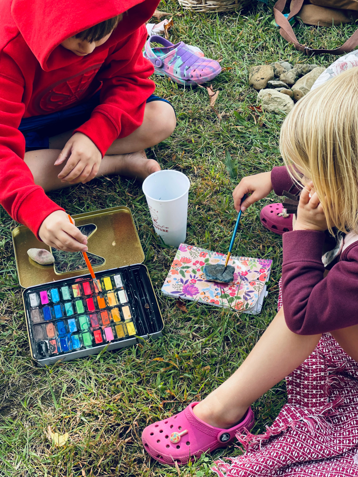 two homeschooled kids sitting on grass painting rocks with water colors.