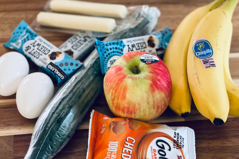 apple, bananas, eggs, cheese sticks, granola bars and goldfish in pile on table.