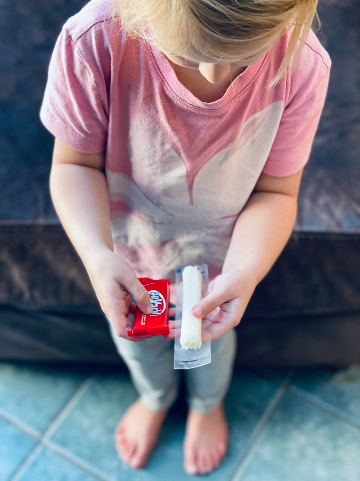 young girl standing on tiled floor, holding a kitkat bar and a cheese stick in hand, trying to decide.