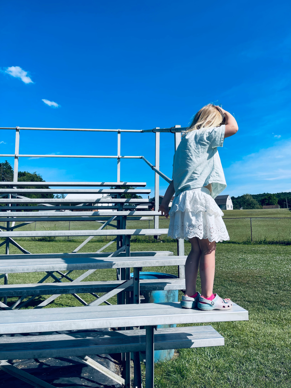 young child standing on bleachers looking down to jump with blue sky in background.