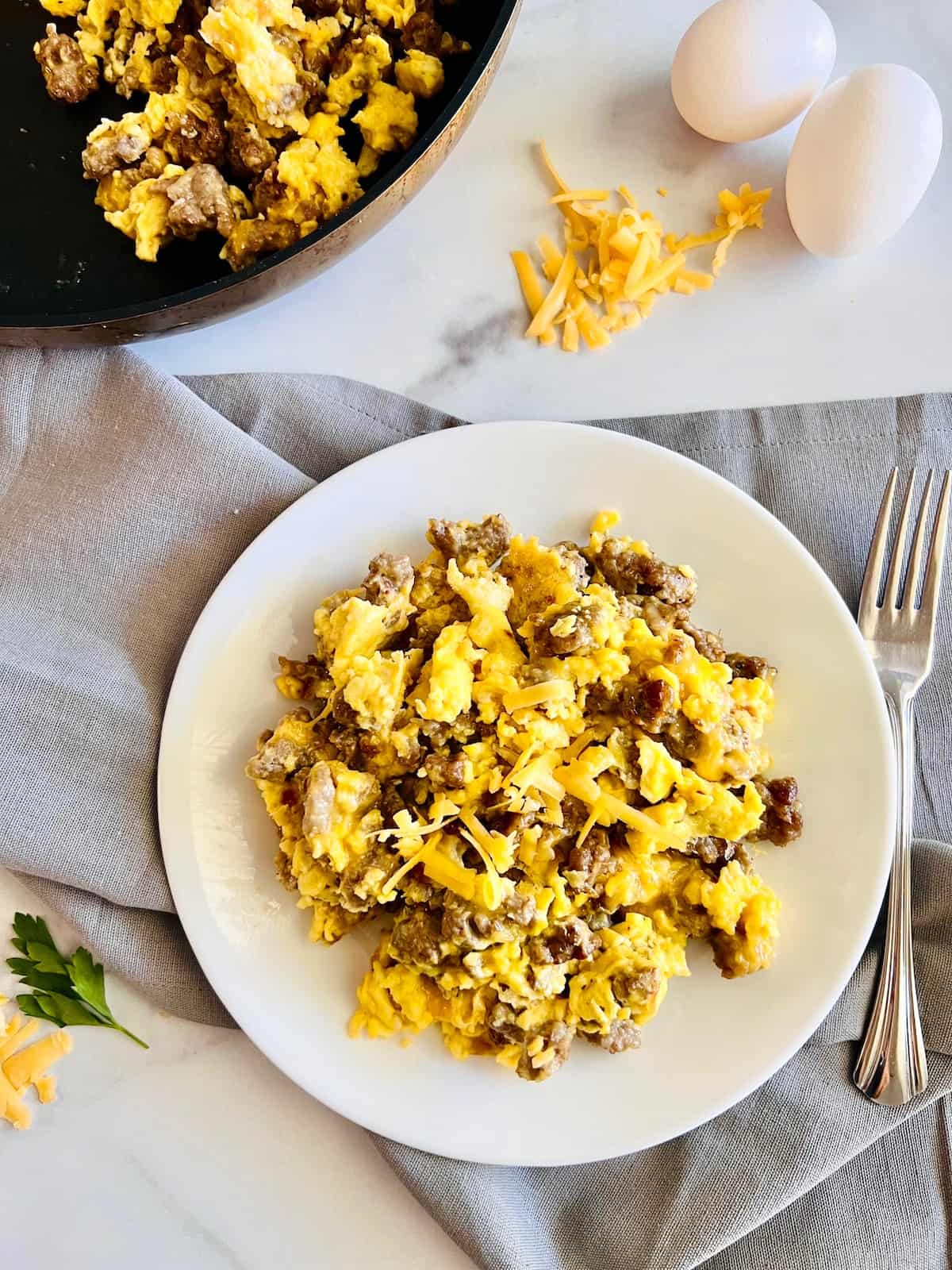 sausage egg and cheese scramble on white plate with fork and raw eggs in background.