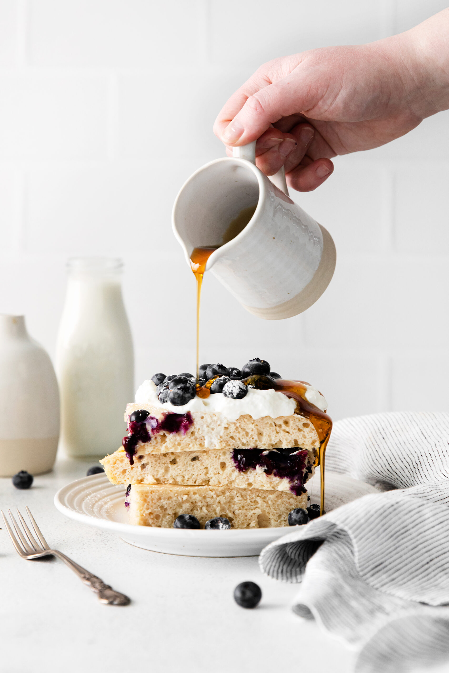 sheet pan pancakes cut and stacked with blueberries and maple syrup being poured.