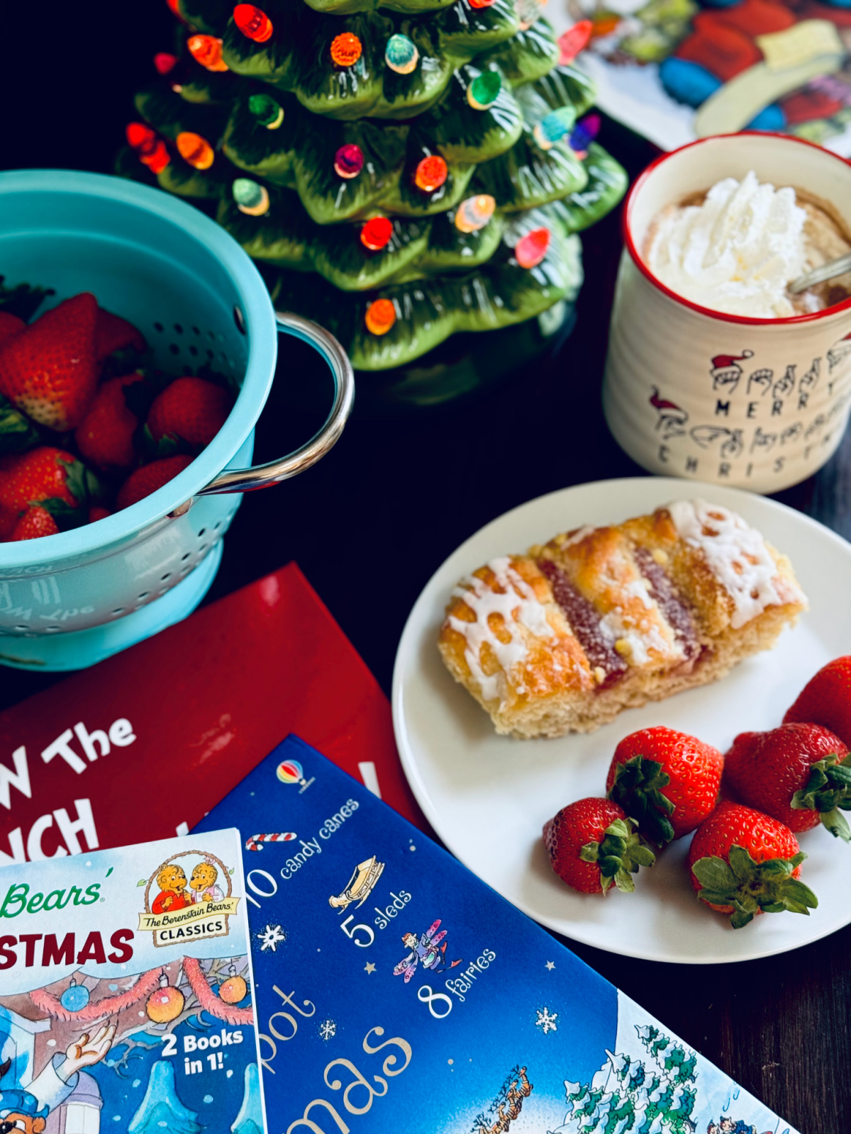 danish and strawberries on white plate with hot chocolate and whipped cream with Christmas picture books and lit ceramic tree in background.