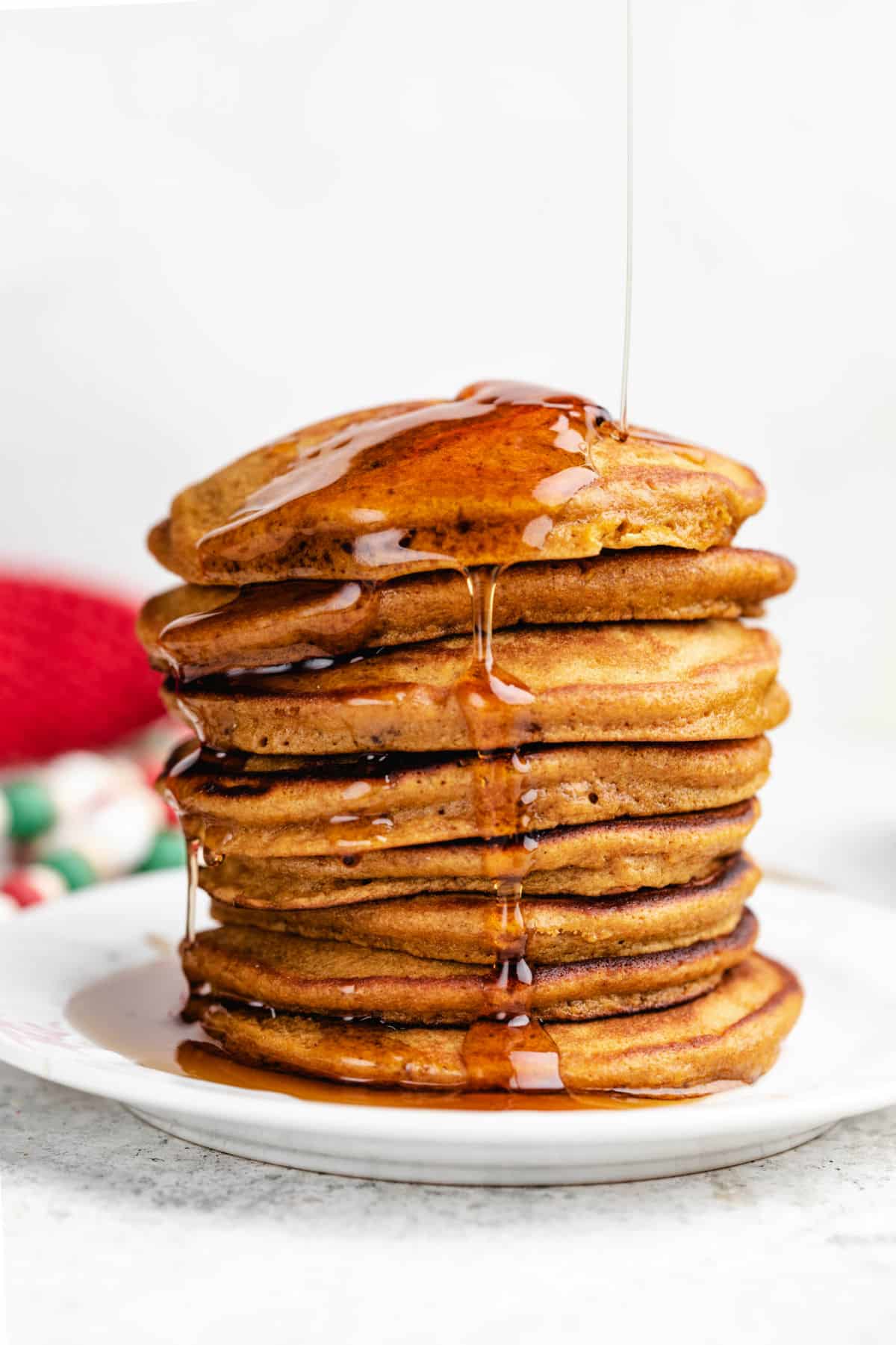 Stack of gingerbread pancakes on a white plate with syrup being poured onto them.