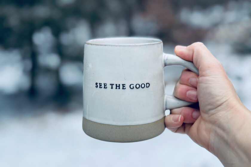 hand holding coffee mug with words, "See The Good" against snowy backdrop.