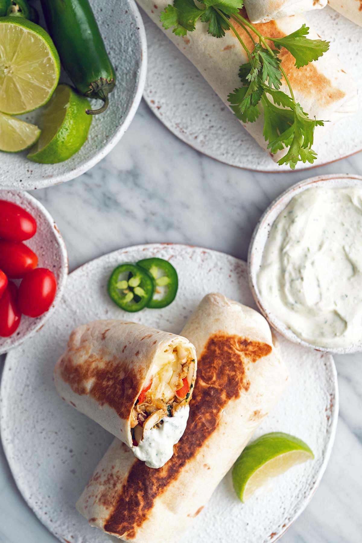 chicken burrito cut in half on plate, surrounded by limes, crema, tomatoes and jalapenos.