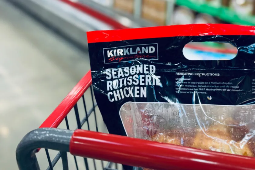 Kirkland Costco rotisserie chicken in bag sitting in grocery cart with red handle.