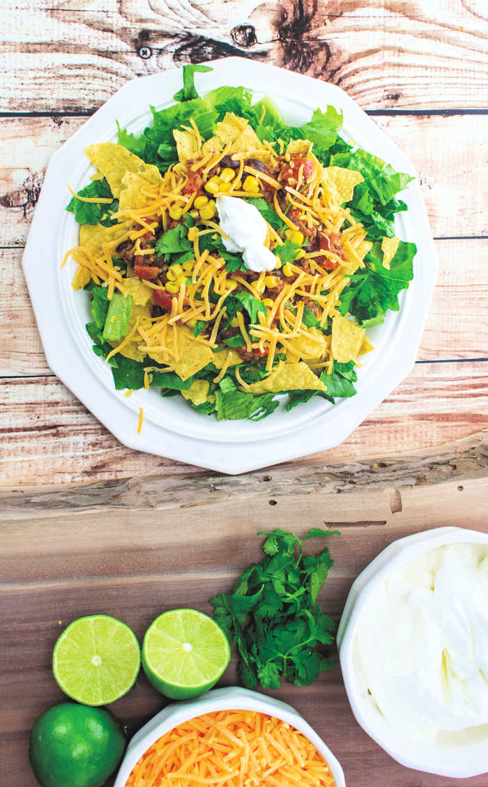 taco salad on white plate with limes, cheese, cilantro and sour cream nearby.