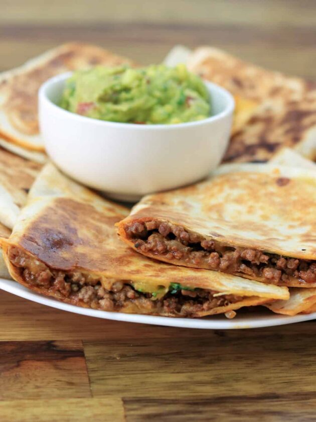 Cheesy ground beef quesadillas cut in slices, layered on plate with small white bowl of guacamole.