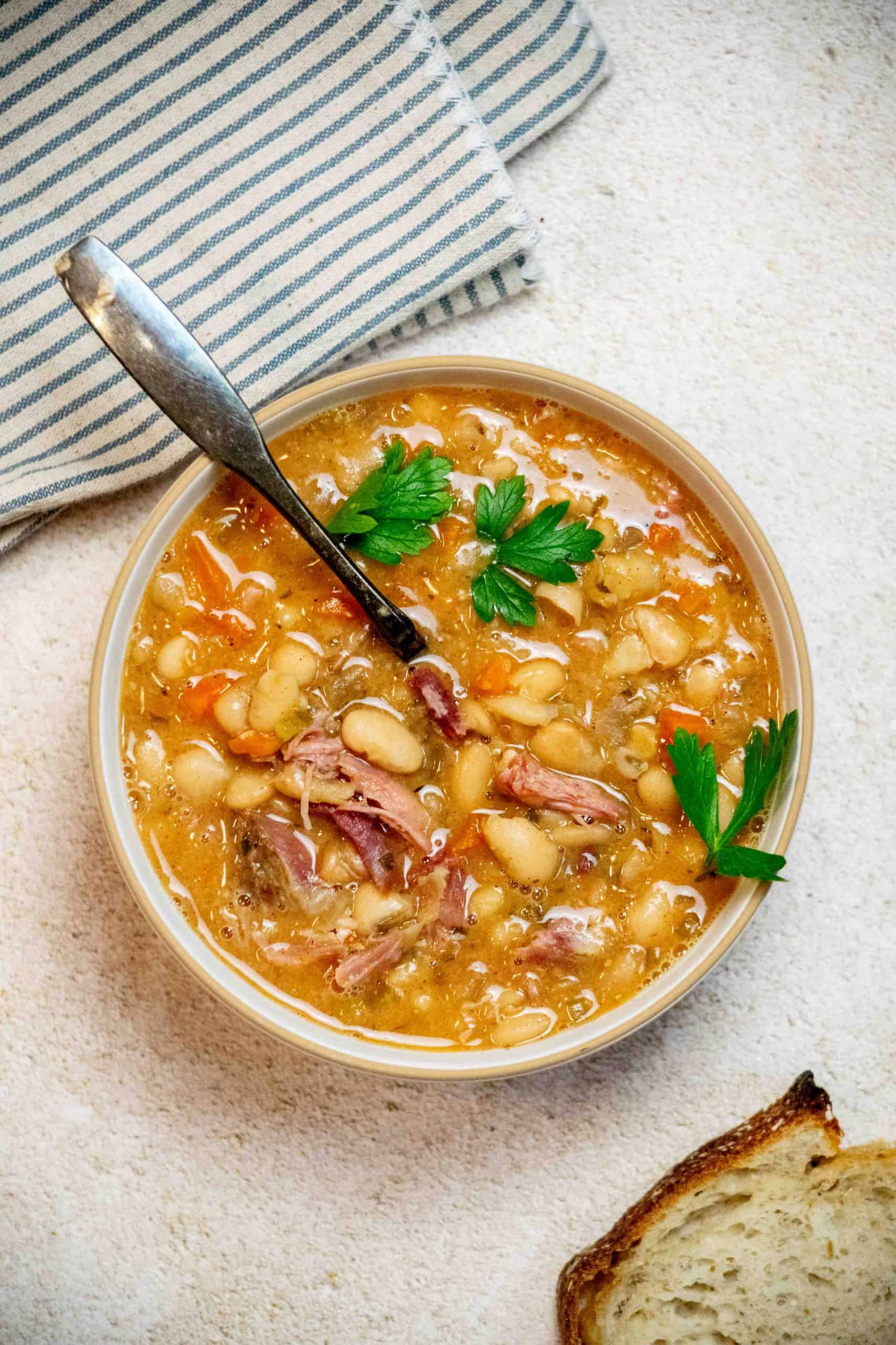 ham bone bean soup in bowl with cilantro leaves and bread slice nearby.