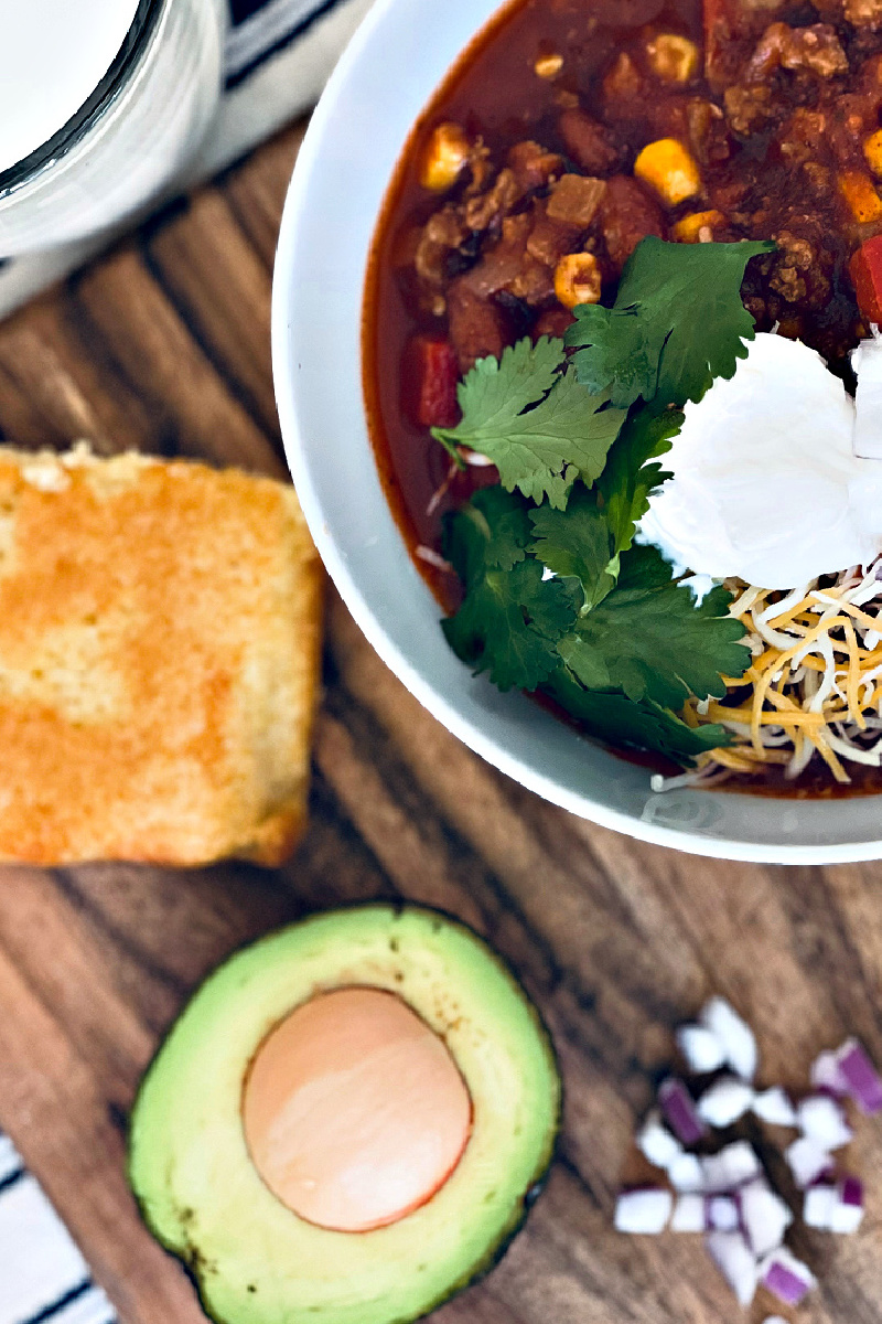 turkey chili in bowl with cilantro and sour cream, with cornbread, avocado and red onion nearby.