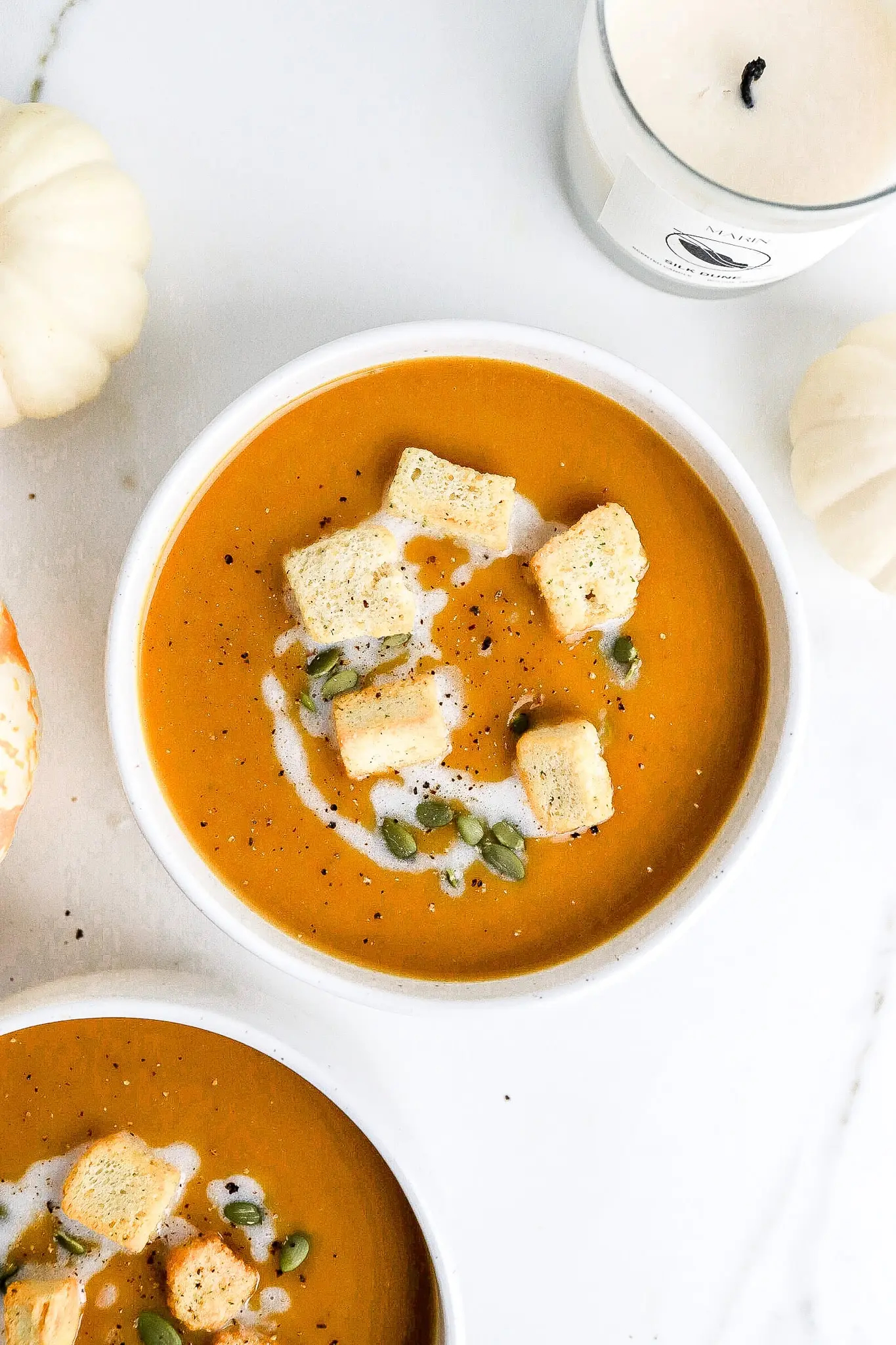 butternut squash soup in white bowl topped with croutons, with white pumpkins and candles nearby.