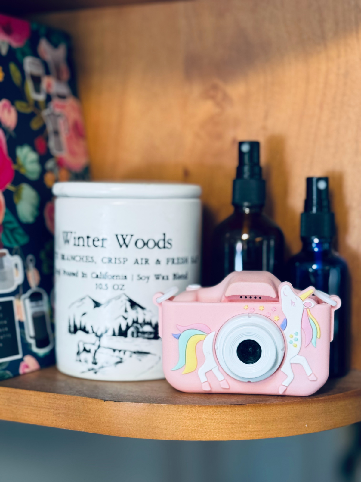 Kids' pink unicorn digital camera in focus with candle jar and spray bottles in background.