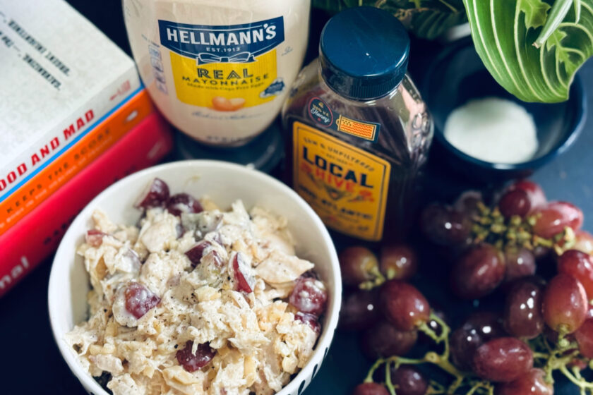 chicken salad in bowl with honey, mayonnaise, books, and grapes in background.