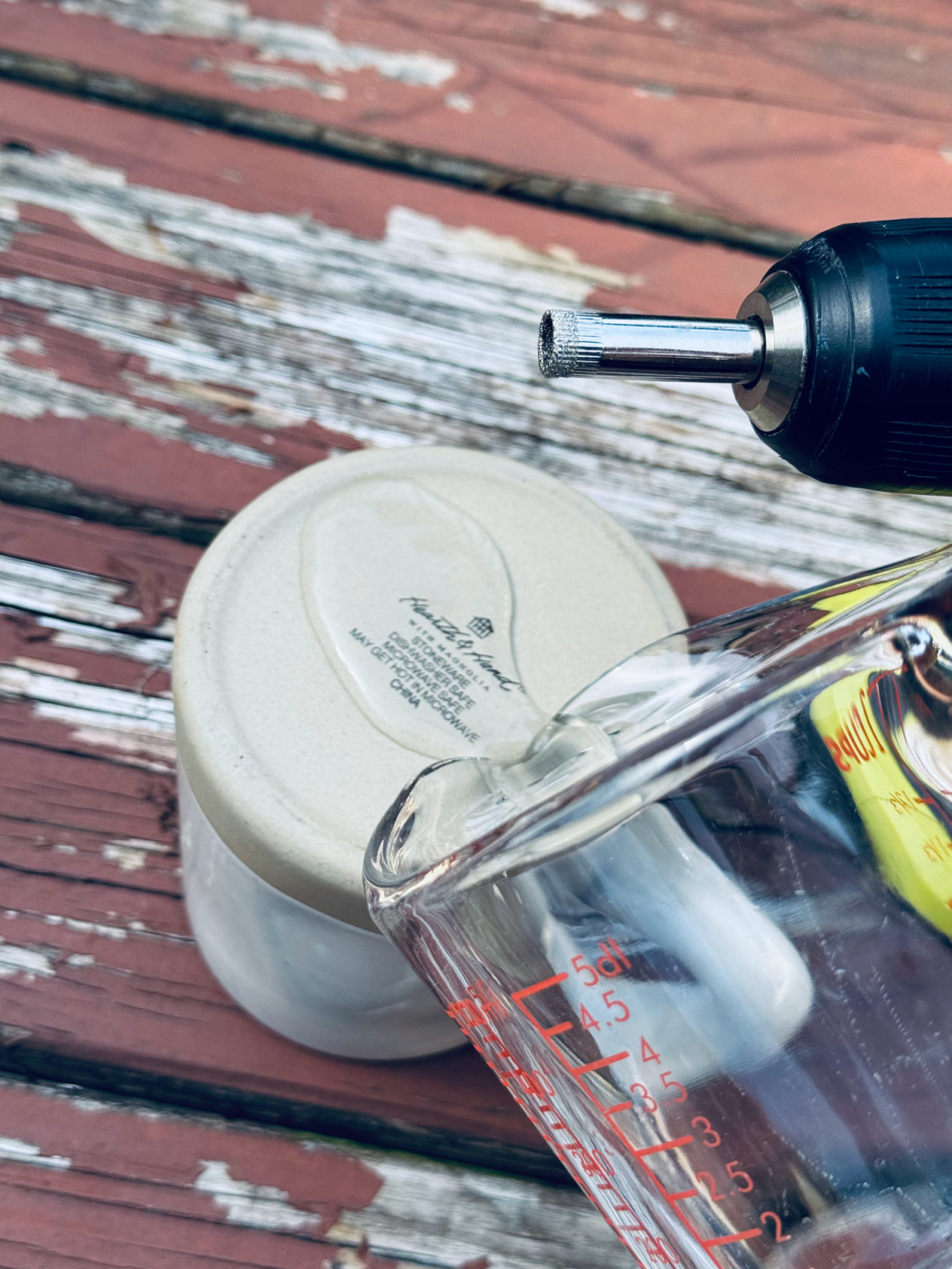 old mug turned upside down on deck, with glass pyrex pouring water onto the bottom, drill with diamond drill bit nearby.
