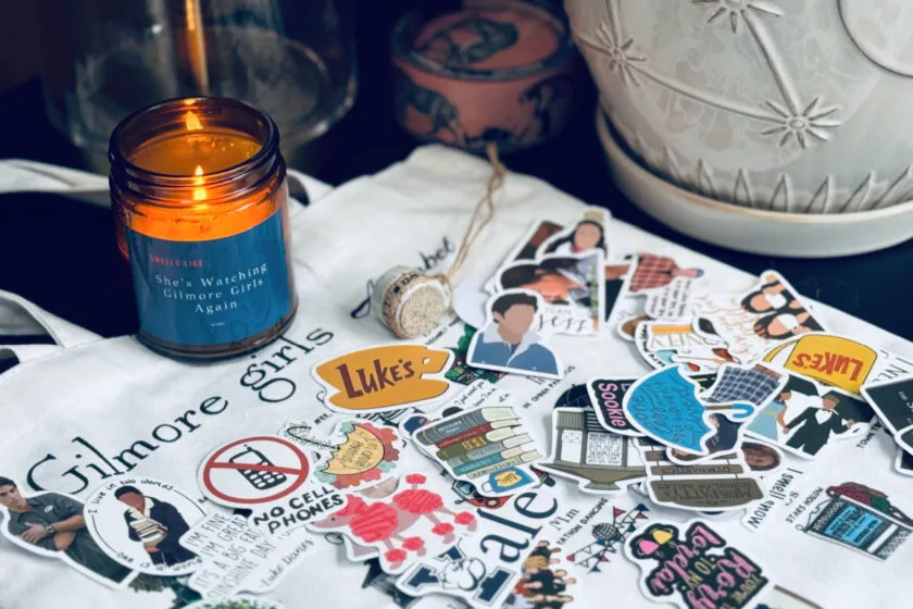 Gilmore Girls stickers and a burning Gilmore Girls candle on top of a Gilmore Girls tote bag.