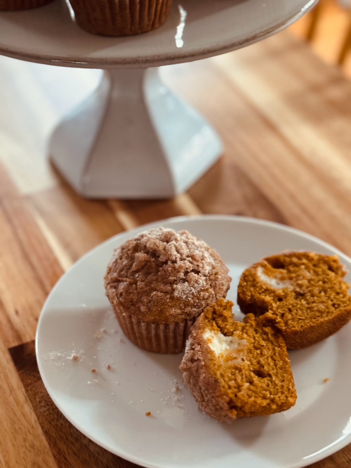 pumpkin cream cheese muffin on white plate, one whole and one cut in half to show cream cheese filling with cake stand in background.