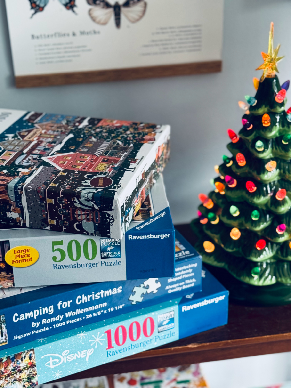 Christmas puzzles stacked on shelf next to lit up ceramic Christmas tree.