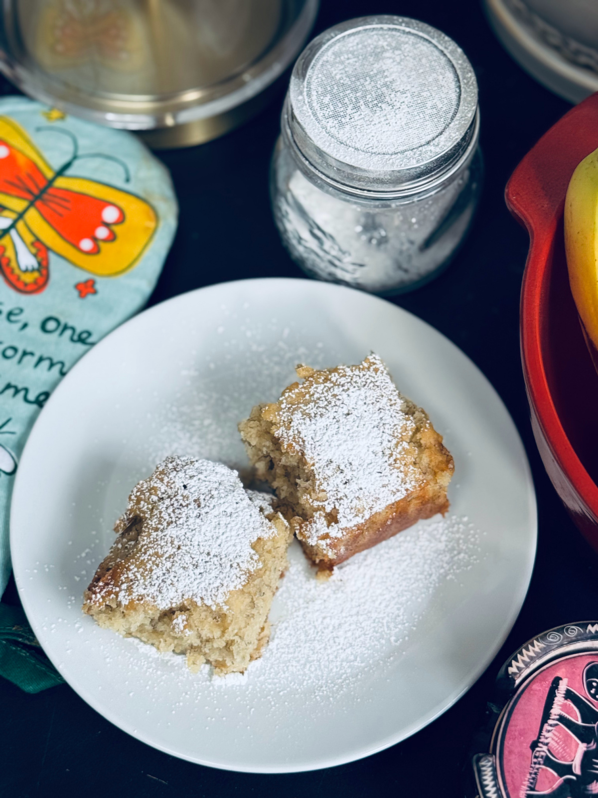 slices of powdered sugared banana bread on white plate with powdered sugar sifter nearby. 