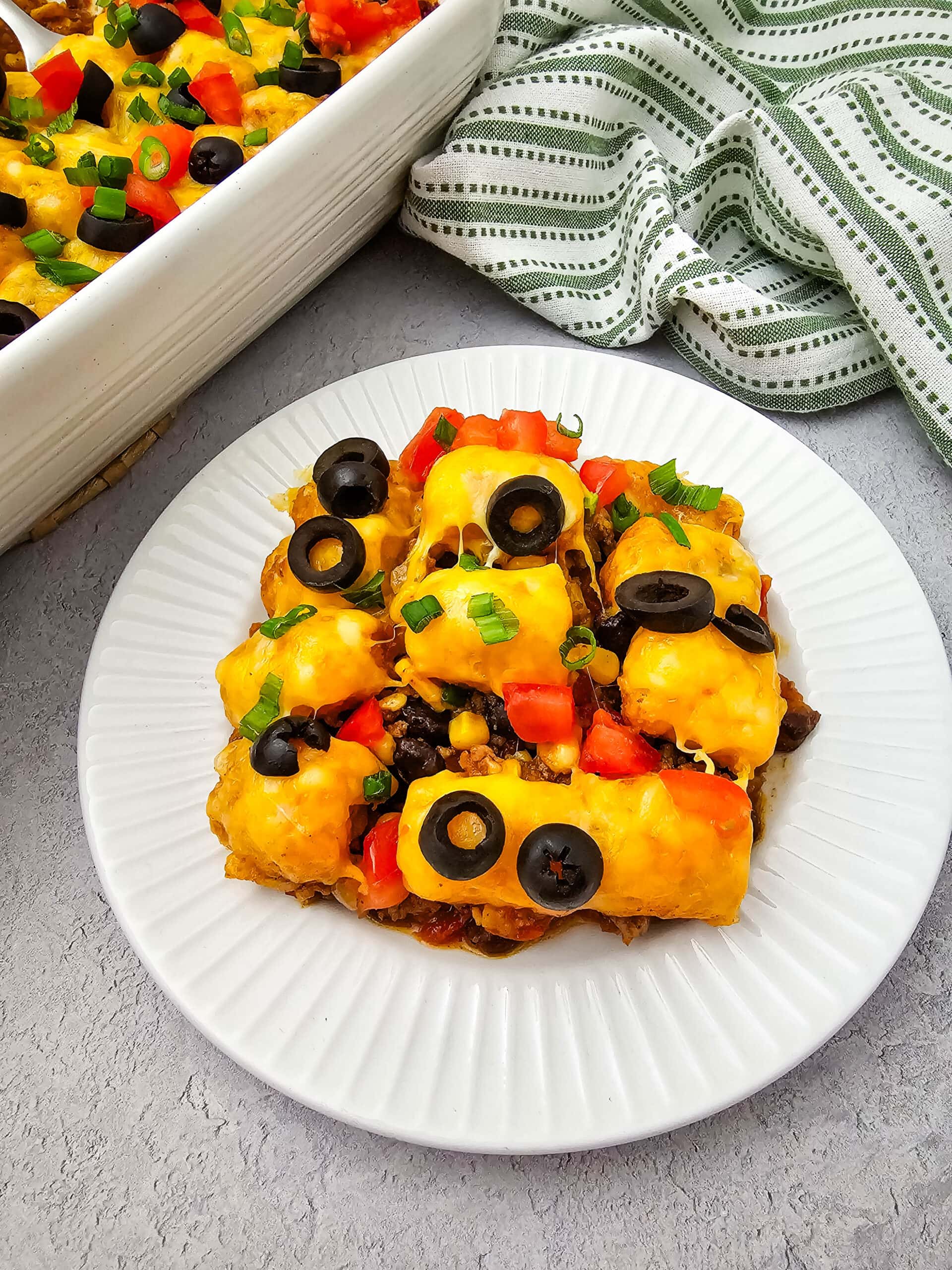 serving of Mexican Taco Tater Tot Casserole on white plate with dish cloth and casserole dish in background.