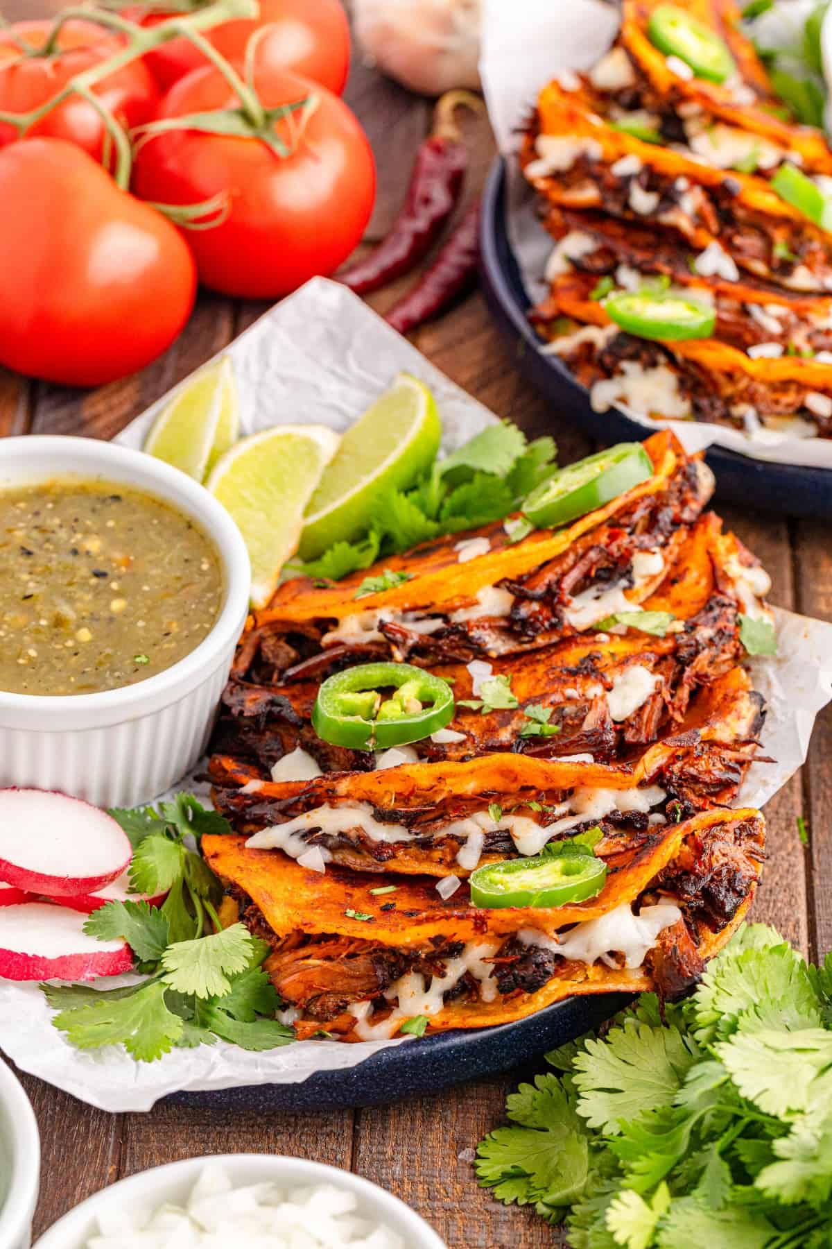 quesabirria tacos prepared on plate, with tomatoes and cilantro in background.