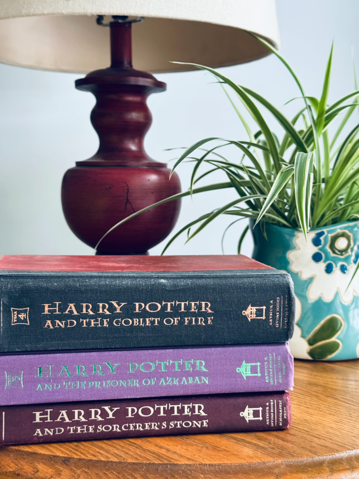 Harry Potter books stacked on table with plant and lamp in background.